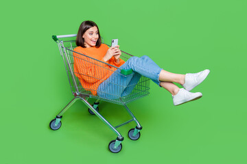 Full size portrait of impressed cheerful girl use smart phone inside hypermarket trolley isolated on green color background - obrazy, fototapety, plakaty