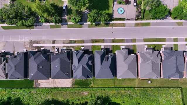Aerial rightward drone tracking view above suburban neighborhood with uniform houses, asphalt shingle roofs, lush backyards, and a clear road with parked cars.