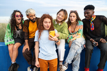 Obraz premium A group of young Gen z millennial friends are sitting on a blue wall looking at a cell phone 