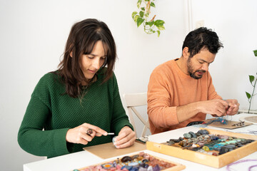 Couple making crafts with stones and macramé. Woman in green sweater filing gem and man weaving bracelet in salmon orange sweater.