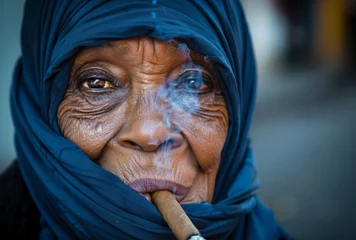 Zelfklevend Fotobehang A woman in a blue scarf is smoking a cigar. She has a sad expression on her face © Sascha