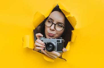 A paparazzi girl in glasses with a rare SLR camera looks out from her hiding place and carefully looks at what is happening. Yellow paper, torn hole. Tabloid press.Looking for a story for stock photos - obrazy, fototapety, plakaty
