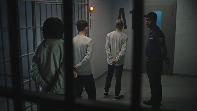 Group of prisoners stand near jail cell, warden talks with them. Multi ethnic teenagers serve imprisonment term in detention center. Young criminals in prison. Justice system. View through metal bars.