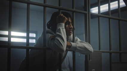 Upset African American teenager with face tattoos stands in prison cell in jail or youth detention center leaning on metal bars. Prison officer passes by young criminal or prisoner in the foreground.