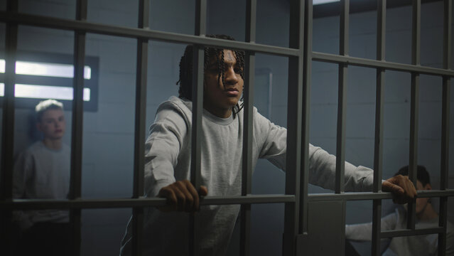 Multiethnic teenagers behind metal bars in prison cell look at camera. Young prisoners, criminals serve imprisonment terms for crimes in jail. Youth detention center or correctional facility. Portrait