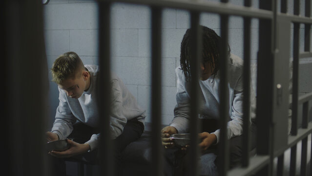 Two multiethnic teenage prisoners sit on bed in prison cell and eat food from iron bowls. Young criminals serve imprisonment term for crimes in jail. Youth detention center or correctional facility.