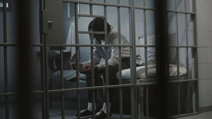 African American young prisoner eats disgusting prison food from iron bowl sitting on bed in prison...