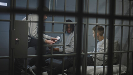 Three multiethnic teenage prisoners play cards in prison cell. Young criminals serve imprisonment term for crimes in jail. Juvenile detention center or correctional facility. View through metal bars.