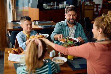Happy family passing food while eating breakfast in hotel.