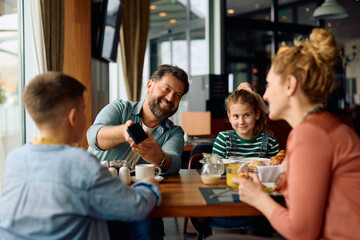 Happy man using smart phone during family breakfast in hotel restaurant. - obrazy, fototapety, plakaty