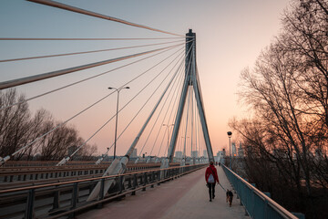 Wide-angle perspective of a suspension bridge at sunset and a person walking a dog