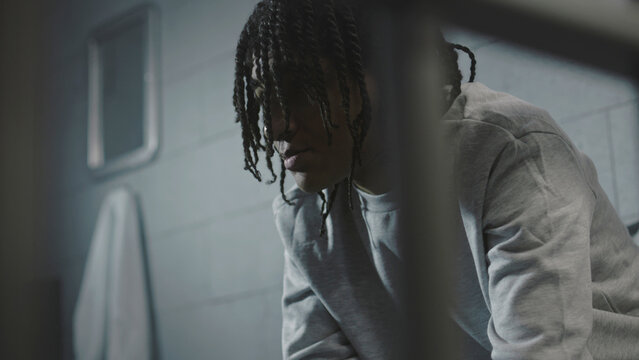 Close up of African American guy trying to eat disgusting prison food sitting on the bed in prison cell in jail. Teenage inmate or young criminal in youth detention center or correctional facility.