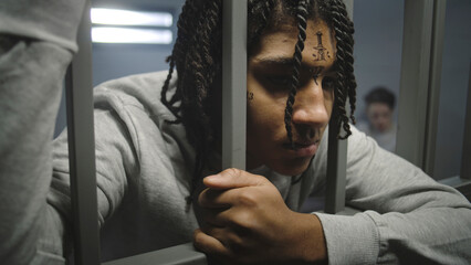 Close up shot of angry African American teenage prisoner with face tattoos standing in prison cell, holding metal bars. Young inmate serves imprisonment term for crime in jail. Youth detention center. - obrazy, fototapety, plakaty