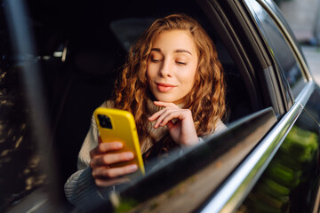 Happy  woman uses a smartphone while sitting in the back seat of a car. Young woman checks mail,...