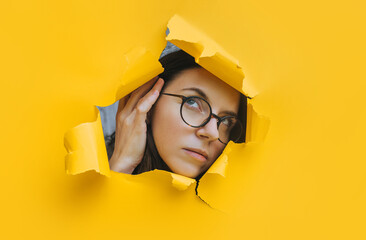 Portrait of a young woman with glasses looking through a hole in yellow paper. An incredulous look. Women's curiosity and gossip. Jealous wife.