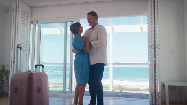 Low angle shot of mature couple with luggage arriving for holiday in beach front property overlooking ocean dancing together - shot in slow motion