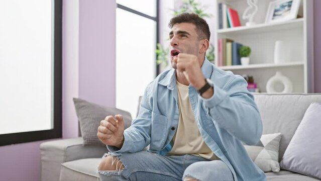 Lucky young hispanic man, bursting with joy, celebrates watching a winning soccer match on tv, crossing fingers in his cozy living room at home