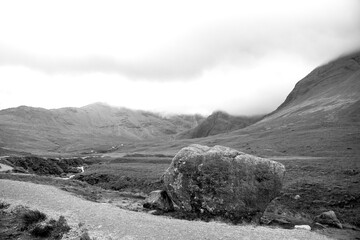 A black and white image of a large boulder along the path to Fairy Pools in Isle of Skye. Misty mountains appear in the background.