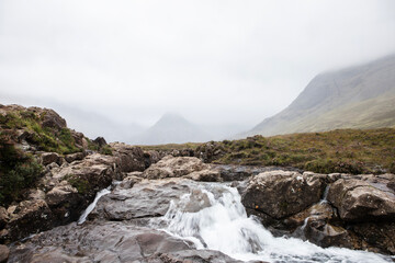 Water flows along a rocky stream. The foreground has mountains covered in fog.