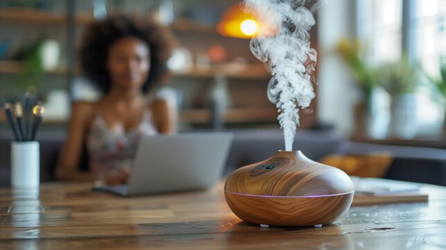 An essential aroma oil diffuser on the table in the office, behind out of focus an African girl is sitting with a laptop on sofa. Concept aromatherapy and relaxing. Air freshener. 