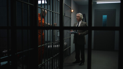 Criminal in orange uniform leans on prison cell bars, talks with advocate and reads lawyer...
