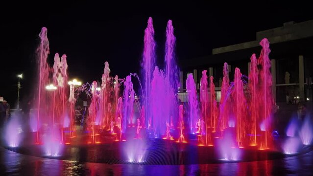 A water feature in the city is illuminated with purple and red lights at night