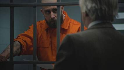 Prisoner in orange uniform leans on prison cell bars, talks with advocate and reads lawyer...