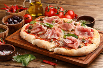 Flatbread pizza with prosciutto, capers and fresh basil leaves on wooden background with ingredients for cooking. Side view, close up, selective focus. Italian food concept