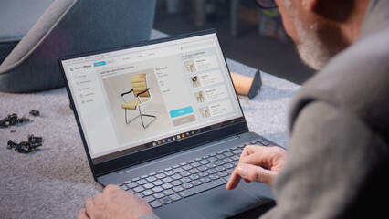 Craftsman uploads pictures of stylish wooden chair to online store using laptop computer. Small...