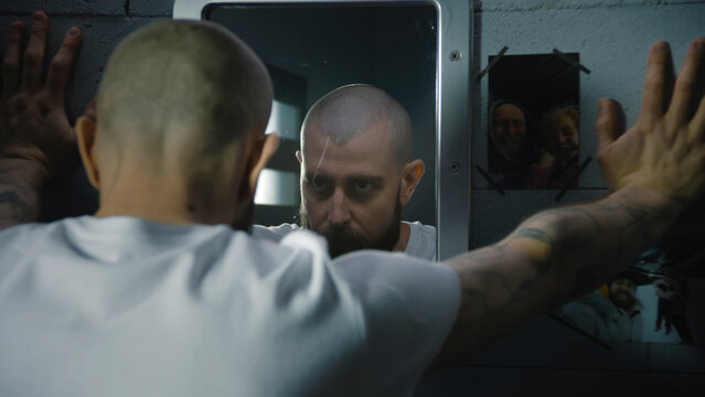 Illegally convicted man looks at himself in the mirror, puts hand on the pictures with family hanging on the wall in prison cell. Male prisoner serves imprisonment term in jail or detention center.