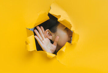 Close-up of a woman's ear and hand through a torn hole in the paper. Bright yellow background, copy space. The concept of eavesdropping, espionage, gossip and tabloids.