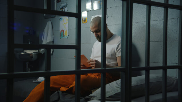 Male prisoner in orange uniform sits on bed in prison cell, looks at photographs of family and children. Criminal serves imprisonment term for crime in jail. Detention center or correctional facility.
