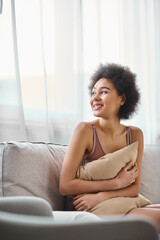 young african american woman with curly hair relaxing on couch in lingerie, smiling and looking away