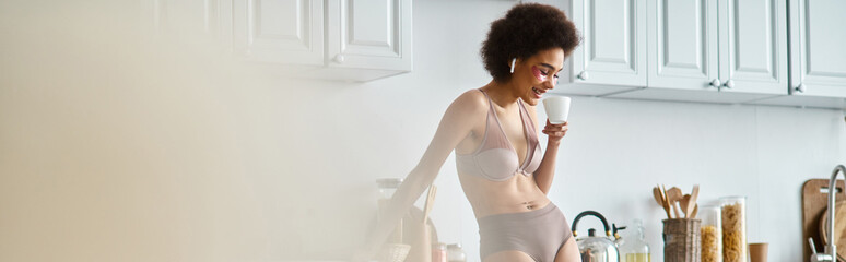 banner of happy african american woman in earphones and lingerie sipping morning coffee in kitchen - obrazy, fototapety, plakaty