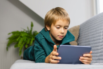 Boy spends time behind a screen on a tablet on the sofa in the living room. Digital device. Technologies.