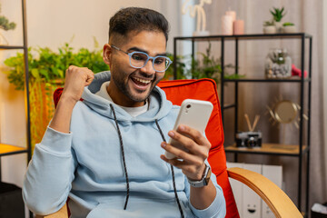 Happy Indian man use mobile smartphone typing browsing say Wow yes, celebrating success victory,...