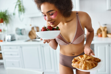 happy black woman in lingerie enjoying smell of strawberries and holding croissants for breakfast - obrazy, fototapety, plakaty