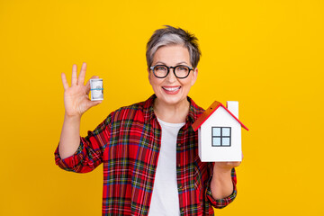 Photo of cheerful grandmother hold rolled stack dollars little house isolated on yellow color background - obrazy, fototapety, plakaty