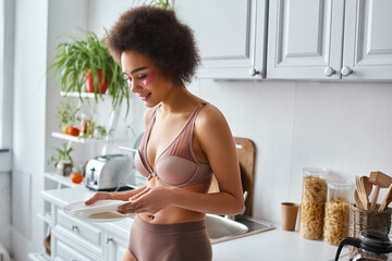 happy and curly african american woman in lingerie with pink patches under eyes holding clean dishes - obrazy, fototapety, plakaty