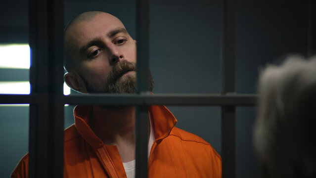 Criminal in orange uniform stands behind bars in prison cell and talks with lawyer. Inmate serves imprisonment term for crime in correctional facility. Prisoner in detention center or jail. Close up.