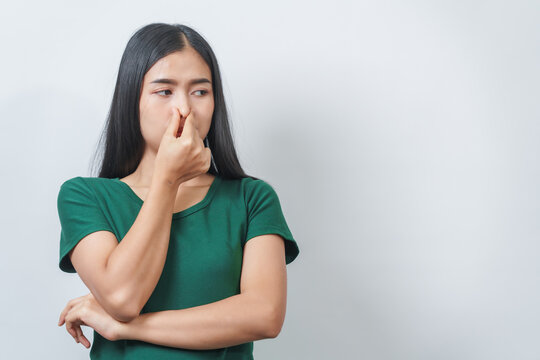 Young Asian frustrated woman with green t-shirt pinches her nose with disgust on her face, reacting to a bad smell in isolation over background.
