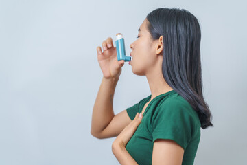 Young Asian woman wearing green t-shirt using asthma inhaler isolated on white background, indoors,...