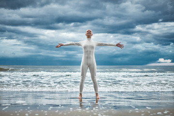 Happy hairless girl with alopecia in white futuristic suit stands with spread arms on beach bathed...