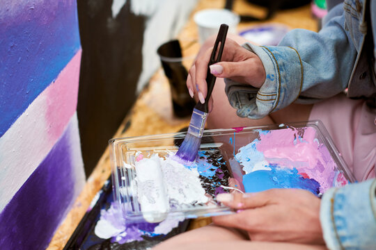 Female painter hand dips paintbrush into palette of colorful paints for live painting of picture for outdoor street exhibition, close up view of female artist hand holding paintbrush