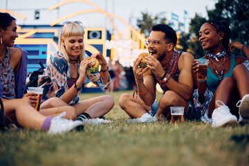 Happy friends eating burgers while attending music festival in summer. - obrazy, fototapety, plakaty