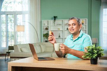 Indian Mid age man working on laptop, smartphone with documents having coffee
