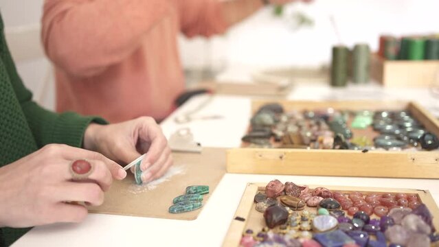 detail of a woman's hand sanding a gemstone, in the background a man weaving bracelets with the gems. Wood with many coloured gems.
