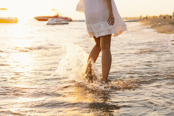 A woman in a dress strolls barefoot along the waters edge, their steps creating ripples in the tranquil sea. Boats float quietly in the background, completing the peaceful scene of an early evening