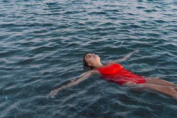 A woman in a vibrant red swimsuit is floating peacefully on her back in the calm waters. Her arms are spread wide, and her eyes are closed in a moment of relaxation as the gentle waves surround her.