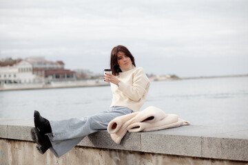 Fototapeta na wymiar A girl with a stylish bob haircut is walking around the city, drinking coffee against the background of water and buildings. The woman is wearing a white faux fur sheepskin coat and jeans